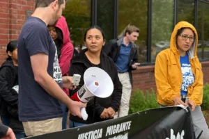 (Wuendy Bernardo, foreground center, a migrant dairy farmworker at risk of deportation, is greeted by Migrant Justice staff member Will Lambek, left, outside a U.S. Immigration and Customs Enforcement office in St. Albans, Vt., on Friday, June 20, 2025. Lambek led a rally in support of Bernardo as she reported to the office for an appointment. (AP Photo: Amanda Swinhart)
