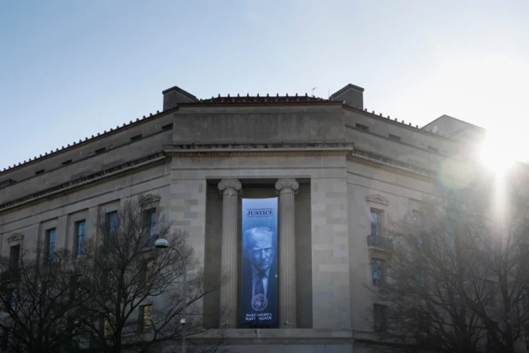 (A banner of President Donald Trump hangs outside the U.S. Department of Justice on Saturday, March 21, 2026, in Washington.  AP Photo: Tom Brenner)