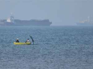 (Two men sit in a small boat on the water as cargo ships are anchored in the background in the Strait of Hormuz off Bandar Abbas, Iran, Monday, May 4, 2026. (Amirhosein Khorgooi/ISNA via AP)