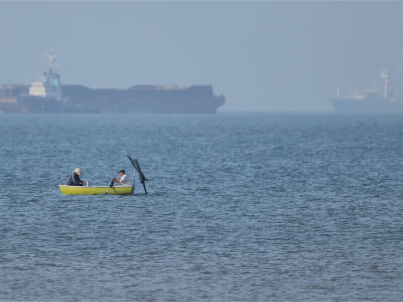 (Two men sit in a small boat on the water as cargo ships are anchored in the background in the Strait of Hormuz off Bandar Abbas, Iran, Monday, May 4, 2026. (Amirhosein Khorgooi/ISNA via AP)