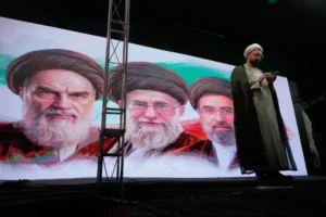 (A cleric checks his cell phone on stage in front of a screen displaying portraits of the late Iranian revolutionary founder Ayatollah Khomeini, left, late Supreme Leader Ayatollah Ali Khamenei, center, and Supreme Leader Ayatollah Mojtaba Khamenei, during a pro-government gathering in Tehran, Iran, Sunday, June 7, 2026.  AP Photo: Vahid Salemi)