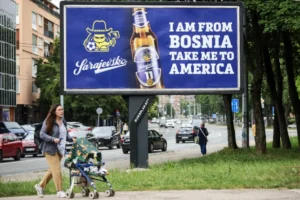 (A woman walks past a billboard displaying lyrics from the Dubioza kolektiv song “I Am From Bosnia, Take Me to America” in Sarajevo, Bosnia, Thursday, June 11, 2026.  AP Photo: Armin Durgut)