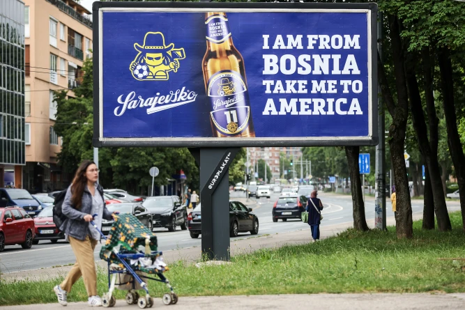 (A woman walks past a billboard displaying lyrics from the Dubioza kolektiv song “I Am From Bosnia, Take Me to America” in Sarajevo, Bosnia, Thursday, June 11, 2026.  AP Photo: Armin Durgut)