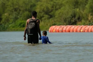(Migrants walk past large buoys being used as a floating border barrier on the Rio Grande, Aug. 1, 2023, in Eagle Pass, Texas. AP File Photo: Eric Gay)
