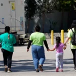 A child holds hands with workers at an emergency shelter for migrant children, July 2, 2021, in Pomona, Calif.  AP File Photo: Marcio Jose Sanchez, Pool)